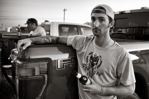 man in t-shirt holds beer with arm on pick-up truck