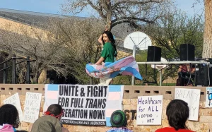 Drag artist Yazmean La Patrona performs on an outdoor stage at Tiguex Park during a Transgender Day of Visibility celebration, waving a transgender pride flag in front of a crowd holding signs supporting trans rights and health care access.