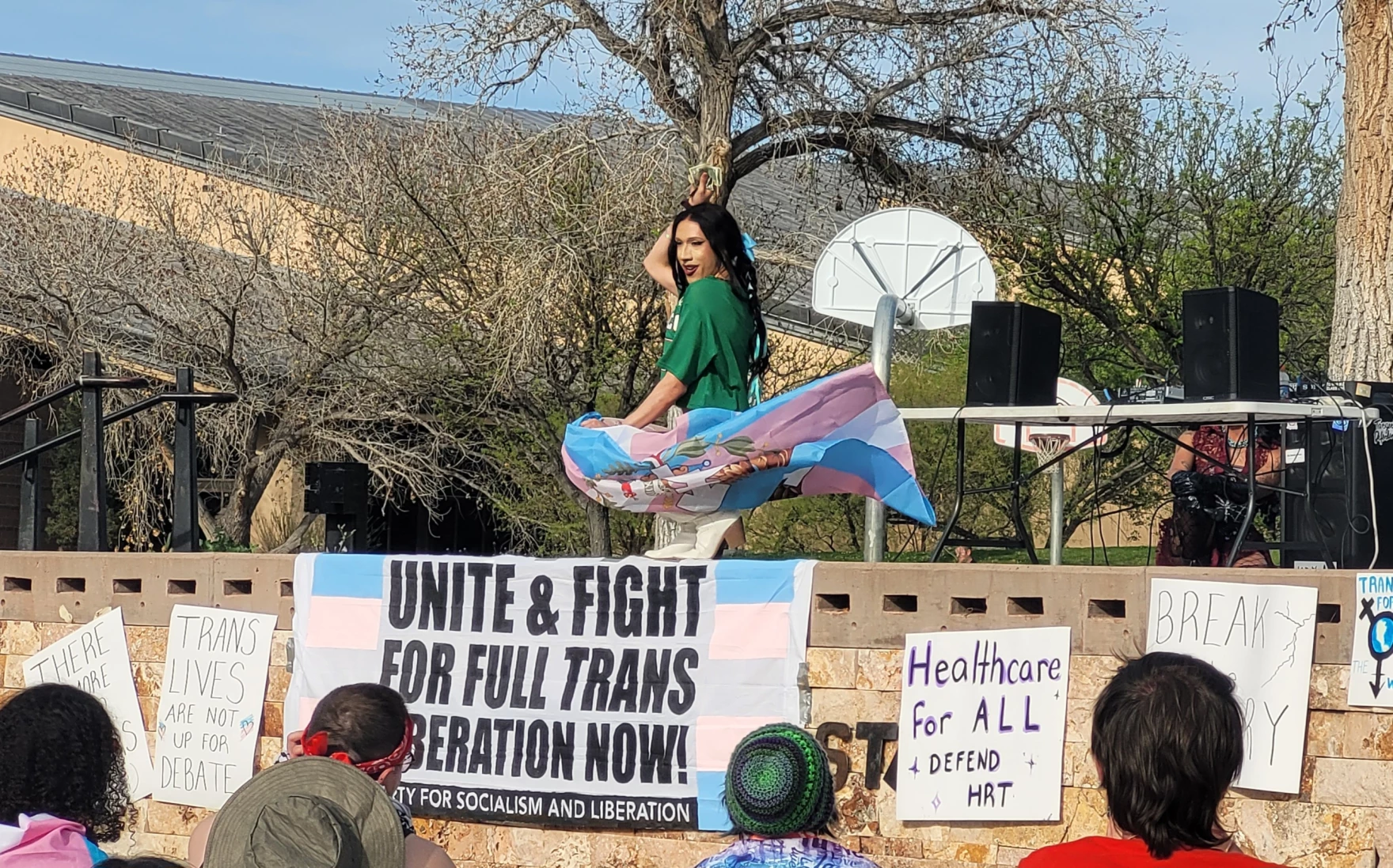Drag artist Yazmean La Patrona performs on an outdoor stage at Tiguex Park during a Transgender Day of Visibility celebration, waving a transgender pride flag in front of a crowd holding signs supporting trans rights and health care access.