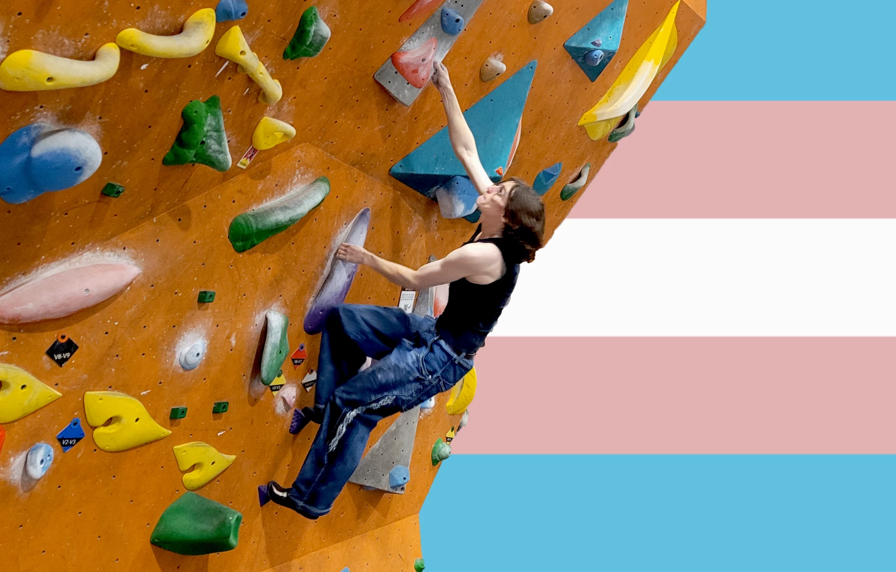 A climber from the group Trans Senders scales an indoor rock wall in front of a large transgender pride flag backdrop, reaching upward toward colorful climbing holds.